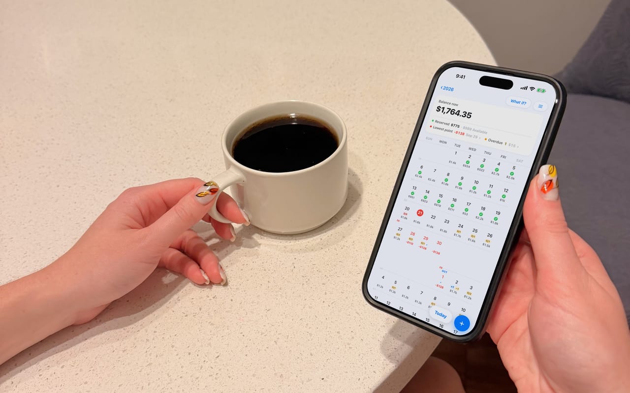 A hand holding an iPhone over a light kitchen table, a cup of black coffee beside it; on the screen, a month in JayCK Flow — every day with its balance, the tight days at the end of the month in red.