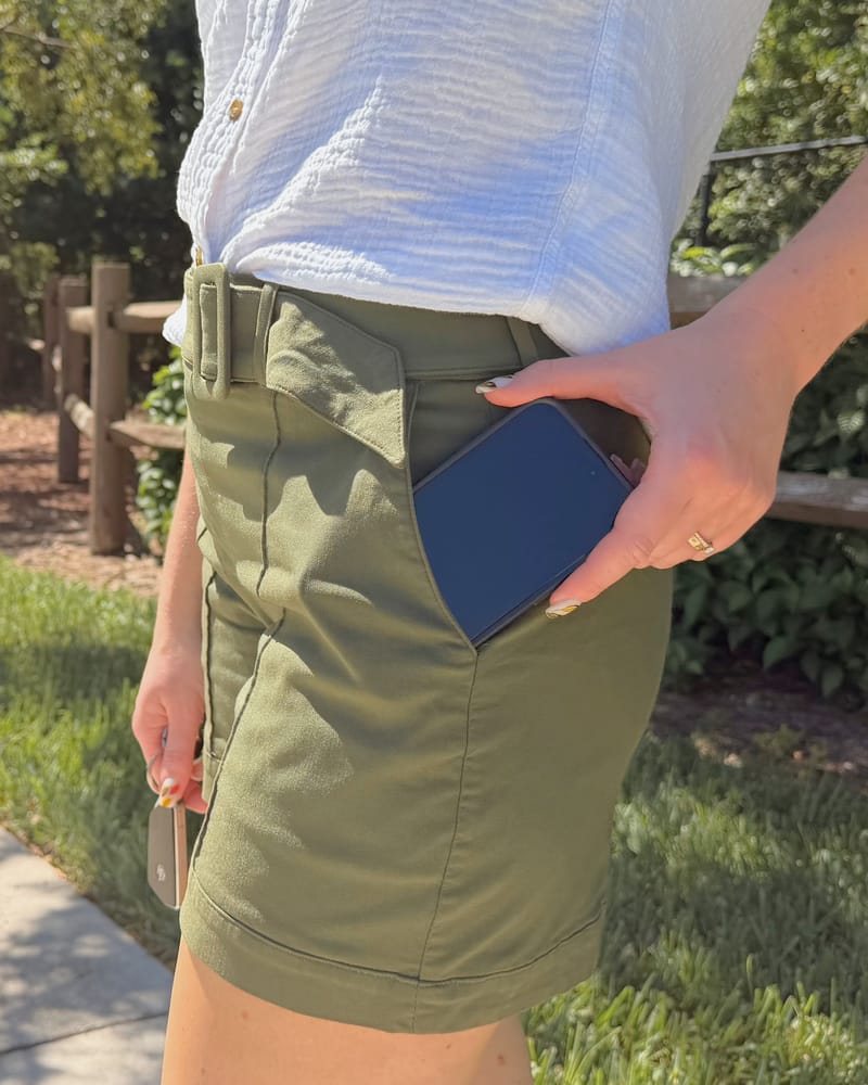 A hand slipping a phone into the pocket of olive shorts, outdoors on a sunny path.