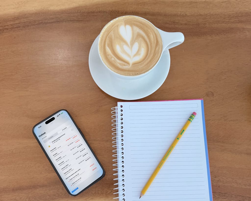 A phone on a wooden table beside a notebook, a pencil and a cup of coffee, seen from above; on the screen, JayCK Flow's outlook — the months ahead, each with its lowest day.
